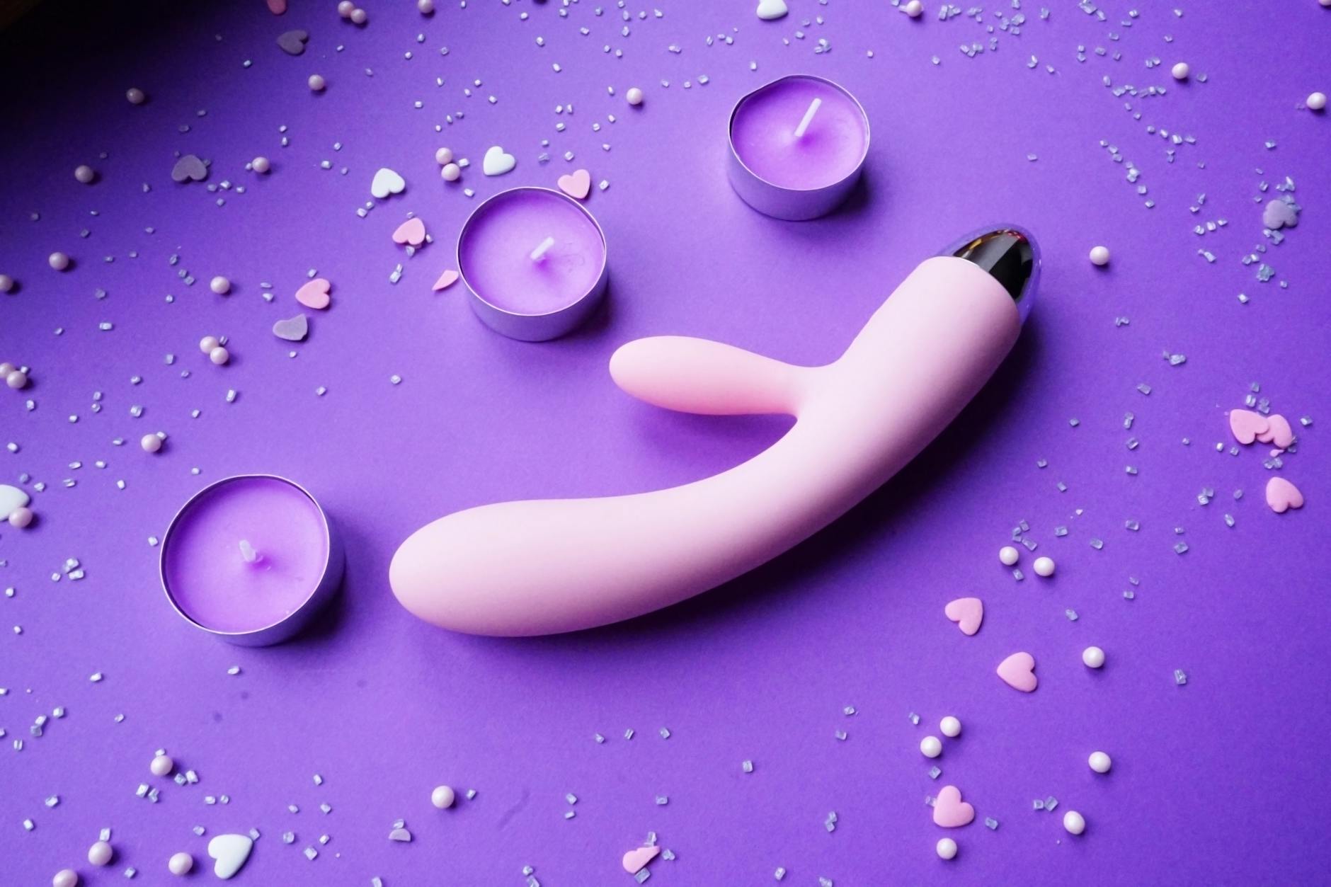 Pink vibrator surrounded by candles and heart confetti on a purple background